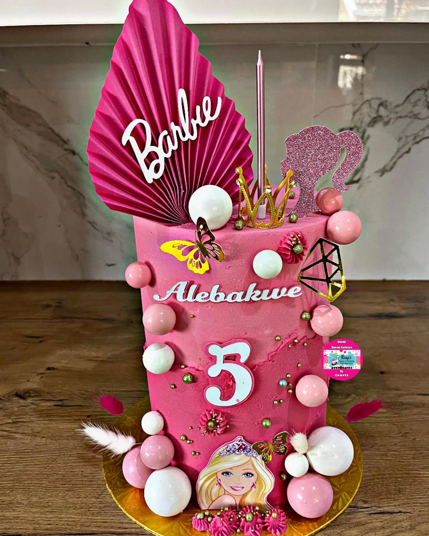 A pink Barbie-themed birthday cake with a gold crown topper, made by Rosy's Cakes and Events
